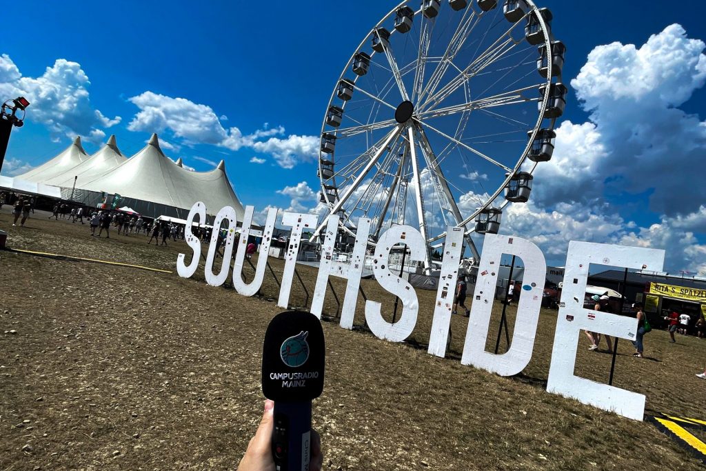 Im Vordergrund ein Aufnahmegerät mit dem Campusradio Mainz Logo, im Hintergrund das Southside Logo auf dem Festivalgelände vor dem Riesenrad.